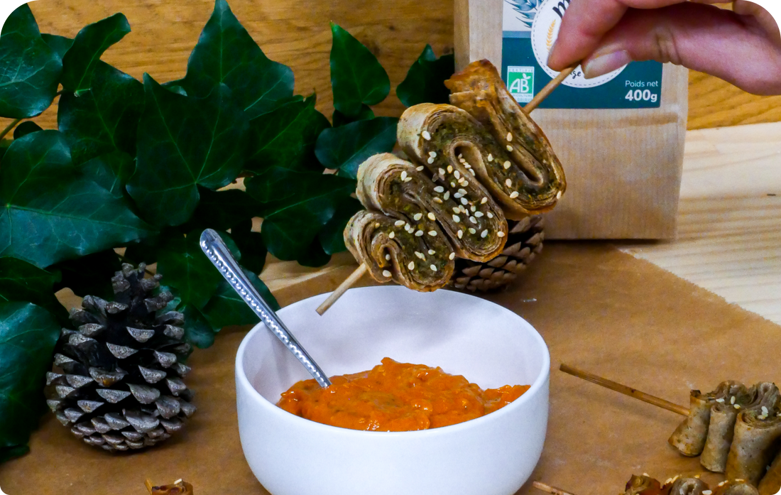 Pâte feuilletée au pesto verde en forme de sapin enfilé sur un pic en bois, qui pointe vers un pot de sauce au pesto rosso sur un fond en bois et de feuilles naturelles