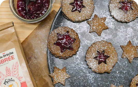 Sablés à la farine de drêche de bière ronds avec des étoiles en confiture au coeur disposés sur une assiettes noir marbrée et entourés d'un pot de confiture et d'un sachet de farine de drêche ambrée